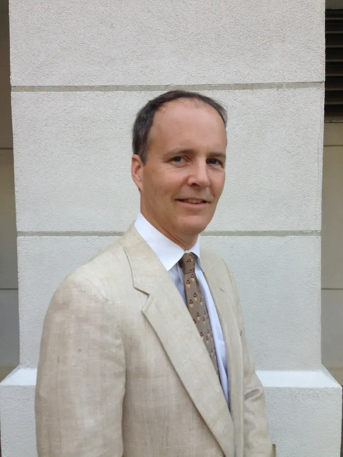 Clifford F. Boyle in a linen jacket and tie, photographed outdoors against a pale limestone wall.